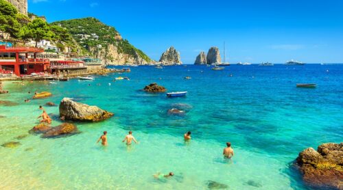 Turistas nadam em praia de Capri com os faragliones ao fundo